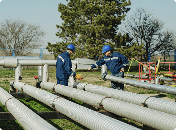 Two individuals standing in front of an industrial setting with large pipes. The men are observing and discussing the infrastructure, creating a scene of collaboration and inspection in an industrial environment.